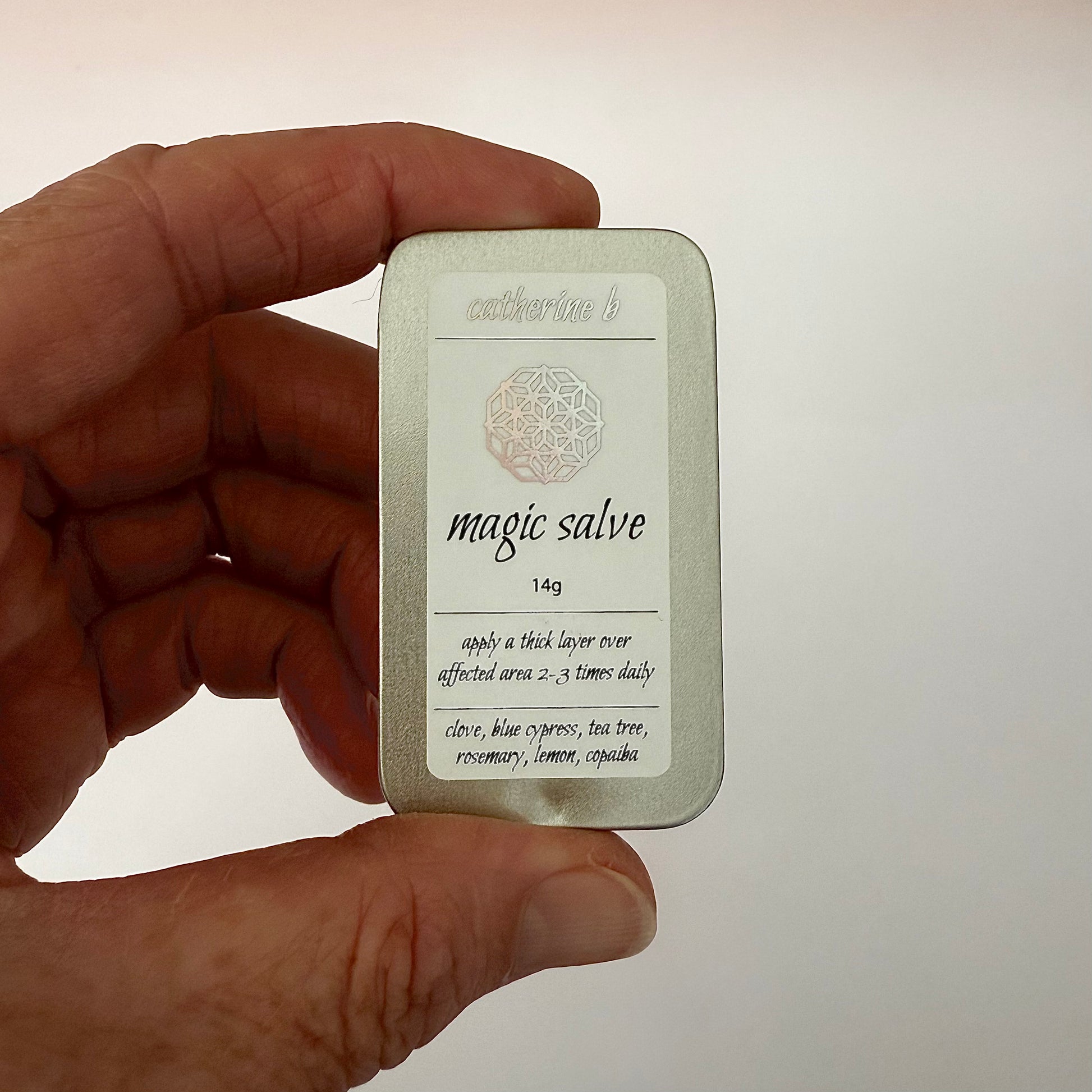Hand holding a medium 14g rectangular tin labeled 'magic salve' with text on a plain background