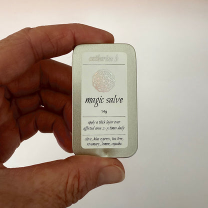 Hand holding a medium 14g rectangular tin labeled 'magic salve' with text on a plain background
