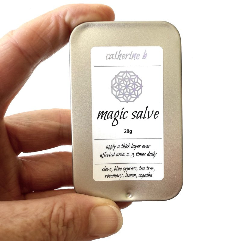 Hand holding a large metal tin labeled 'magic salve' with a hand cream container on a white background.